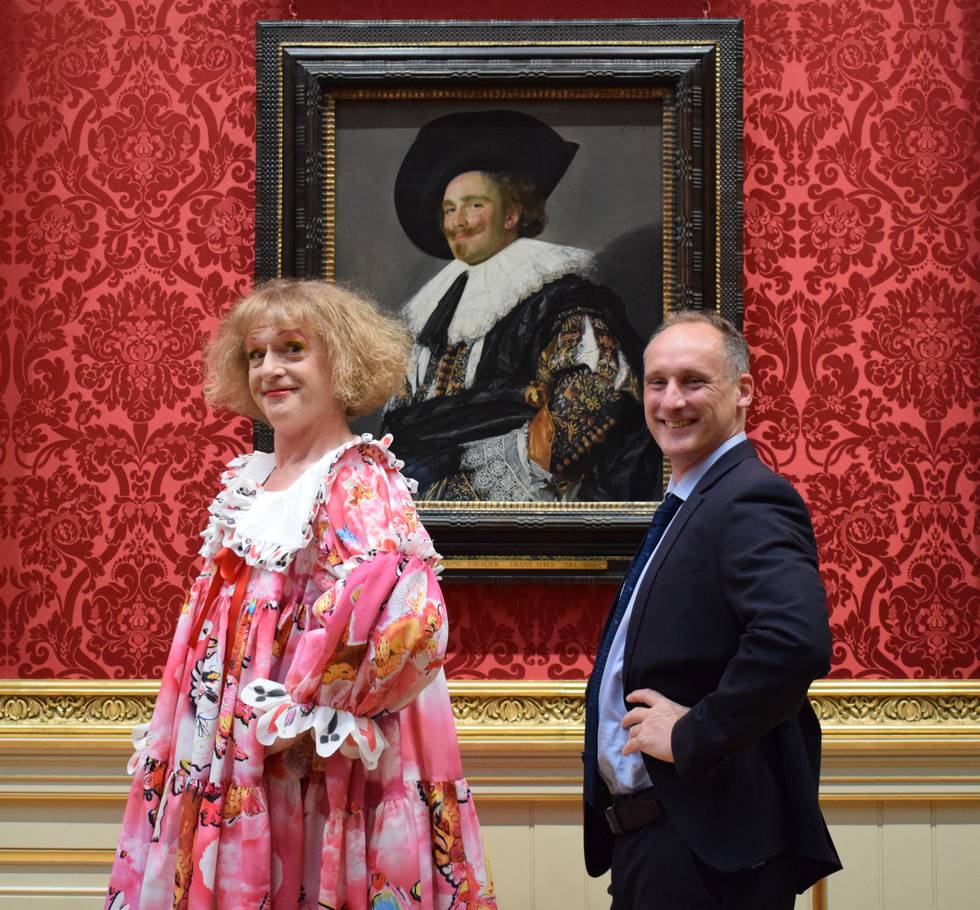 Two people stood in front of a painting of a man with a beard wearing a hat